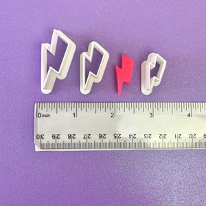 May include: Three white plastic lightning bolt cookie cutters of different sizes on a purple background with a ruler for scale. The smallest cutter is red.