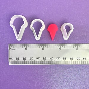 May include: Four white plastic ice cream cone cookie cutters, two large, one medium, and one small, with a red plastic ice cream cone cookie cutter in the middle. The cookie cutters are on a purple surface with a ruler measuring 10 cm.