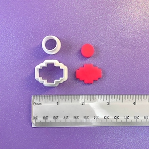 May include: A set of three white plastic cookie cutters in the shape of a circle, a cross, and a square. The cutters are on a purple surface with a ruler for scale. The ruler is marked in inches and 1/16ths of an inch.