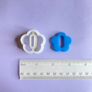May include: Two cookie cutters in the shape of a flower with a number "0" inside. The cutter on the left is white plastic and the cutter on the right is blue plastic. A ruler is below the cutters, showing the size of the cutters to be about 1 inch.