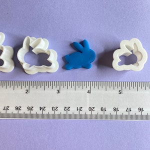 May include: Four white plastic cookie cutters shaped like bunnies, one with a solid blue bunny in the center. The cutters are lined up on a purple surface with a ruler underneath.