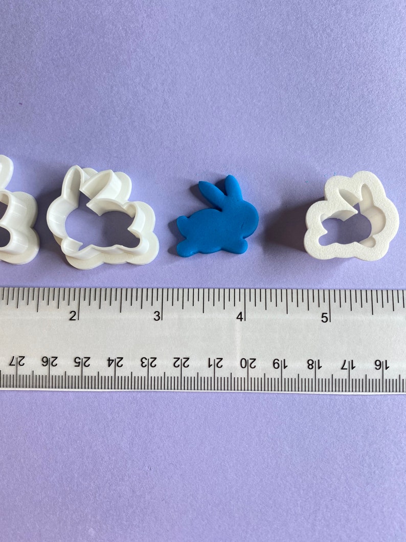 May include: Four white plastic cookie cutters shaped like bunnies, one with a solid blue bunny in the center. The cutters are lined up on a purple surface with a ruler underneath.