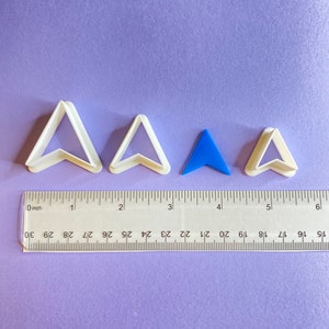 May include: Four cookie cutters in the shape of an arrow. Three are white plastic and one is blue plastic. The cutters are lined up on a purple surface with a ruler for scale.