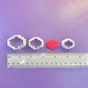 May include: Four cookie cutters, two white and two red, shaped like pixelated hearts. The cutters are arranged in a row on a purple surface with a ruler.