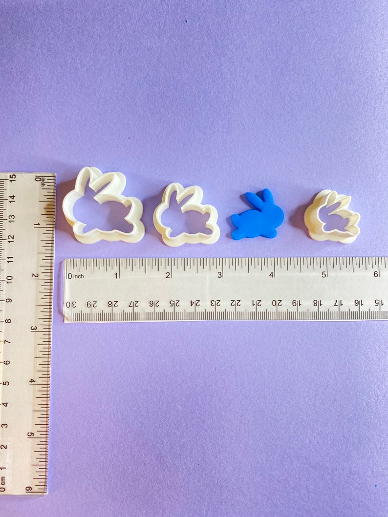 May include: Four white plastic bunny cookie cutters of varying sizes. The largest cutter is approximately 2 inches wide. The smallest cutter is approximately 1 inch wide. The cutters are on a purple surface with a ruler for scale.