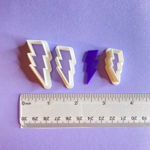 May include: Four cookie cutters in the shape of a lightning bolt. Two are white plastic, one is purple plastic, and one is white plastic with a purple outline. A ruler is in the bottom of the image with markings in inches.