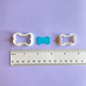 May include: Two white plastic dog bone cookie cutters and a blue plastic dog bone cookie cutter on a purple surface with a ruler measuring 5 inches.