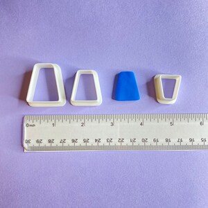 Peut inclure: Trois emporte-pièces en plastique blanc en forme de rectangle, de triangle et de trapèze. Un emporte-pièce en plastique bleu en forme de triangle est au centre. Une règle est placée sous les emporte-pièces, avec le repère 0 pouce visible.