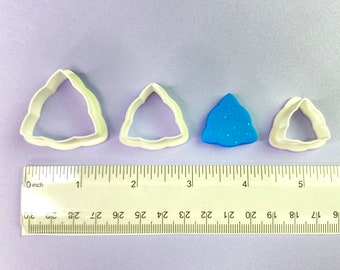 Puede incluir: Tres cortadores de galletas de plástico blanco en forma de triángulo de diferentes tamaños, con un triángulo azul de arcilla brillante en el centro. Una regla se encuentra debajo de los cortadores, con los números del 0 al 5 pulgadas marcados.
