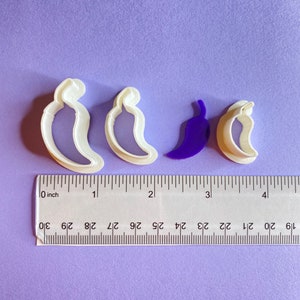 Puede incluir: Cuatro cortadores de galletas con forma de chiles. Dos son blancos, uno es morado y uno es blanco con un contorno morado. Los cortadores están dispuestos en una fila sobre una superficie morada con una regla debajo.