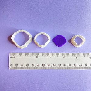 May include: Four white plastic cookie cutters in the shape of seashells, with a purple piece of fondant in the middle. The cutters are lined up from largest to smallest, with a ruler underneath for size reference.