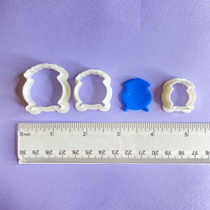 May include: Four white plastic cookie cutters in the shape of cauldrons, with a blue playdough cauldron in the centre. The cutters are arranged in order of size, with the largest on the left and the smallest on the right. A ruler is visible at the bottom of the image.