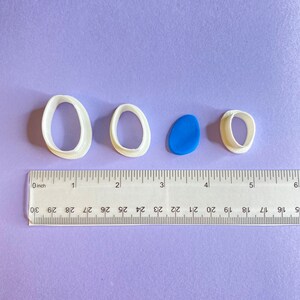 May include: Three white plastic cookie cutters in the shape of ovals, with a blue polymer clay oval in the center. A ruler is visible at the bottom of the image, with markings in inches.
