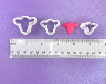 Op de afbeelding: Drie witte plastic koekjesvormen in de vorm van koeien, in verschillende maten. De grootste vorm is 5 cm breed, de middelste vorm is 3,8 cm breed en de kleinste vorm is 2,5 cm breed. De vormen liggen op een paarse ondergrond met een liniaal voor schaal.