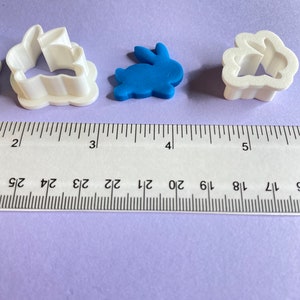 May include: Three cookie cutters, one white bunny, one blue bunny, and one white cloud with a bunny inside. The cutters are on a purple surface with a ruler for scale.