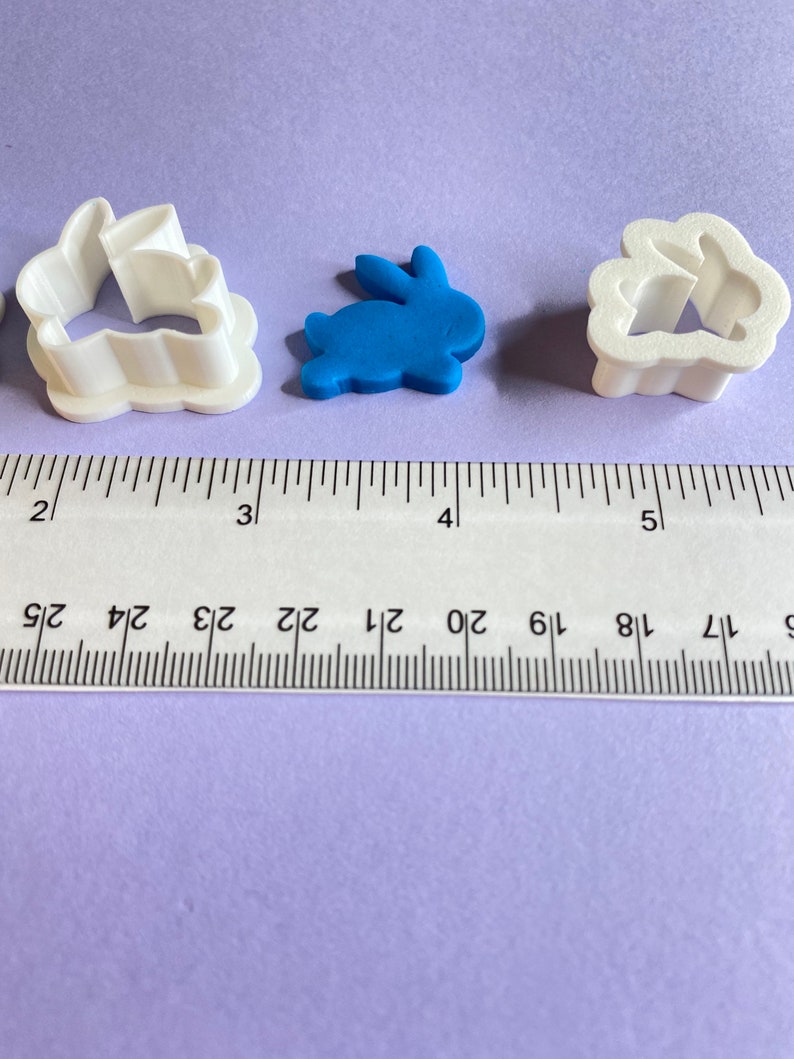 May include: Three cookie cutters, one white bunny, one blue bunny, and one white cloud with a bunny inside. The cutters are on a purple surface with a ruler for scale.