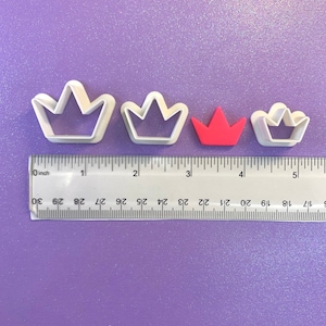 May include: Four white plastic crown cookie cutters of varying sizes, with a red plastic crown cookie cutter in the center. The cookie cutters are arranged in a row on a purple surface with a ruler underneath.
