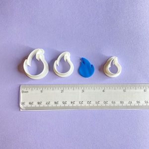 Op de afbeelding: Vier koekjesvormen in de vorm van vlammen. Drie zijn van wit plastic en één van blauw plastic. De vormen zijn in een rij op een paarse ondergrond geplaatst met een liniaal eronder.