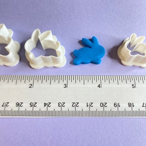 May include: Three white plastic bunny cookie cutters and a blue fondant bunny on a purple surface. A ruler is placed below the cutters, showing the size of the cutters.
