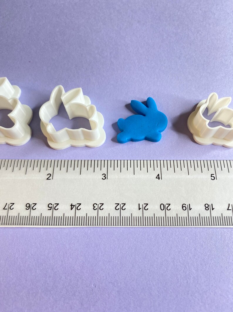 May include: Three white plastic bunny cookie cutters and a blue fondant bunny on a purple surface. A ruler is placed below the cutters, showing the size of the cutters.