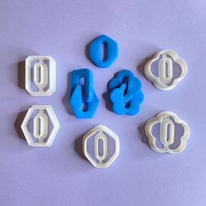 May include: A set of eight cookie cutters, including a hexagon, a flower, and a chain link. The cutters are made of white plastic and are arranged on a purple background. Two of the cutters are blue.