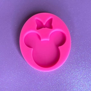 May include: A vibrant pink silicone mould in the shape of a cartoon mouse head with a bow. The mould is oval-shaped and sits on a purple glittery surface. The mouse head has two large ear shapes and a bow at the top.