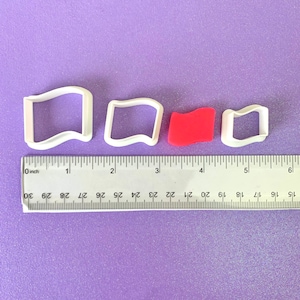 May include: Four white plastic cookie cutters in the shape of a wavy rectangle. The cutters are arranged in a row from largest to smallest. The largest cutter is about 1 1/2 inches wide. The smallest cutter is about 1 inch wide. A red plastic cookie cutter is in the middle of the row. The cutters are on a purple surface with a ruler underneath.