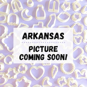May include: A white background with a variety of white cookie cutters in different shapes, including a heart, a star, a lightning bolt, a flower, and a crescent moon. The text "ARKANSAS PICTURE COMING SOON!" is written in black letters on a white rectangle in the center of the image.