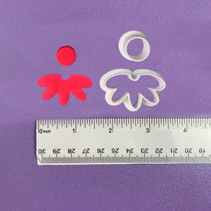 Puede incluir: Dos cortadores de galletas de plástico blanco en forma de flor. El cortador más grande tiene cinco pétalos y un círculo en el centro. El cortador más pequeño tiene cinco pétalos. Un círculo rojo y una galleta con forma de flor roja son visibles en el fondo.