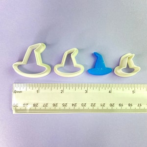 Könnte beinhalten: Vier Ausstecher aus Kunststoff in Form von Hexenhüten. Die Ausstecher sind weiß, bis auf einen, der blau mit Glitzer ist. Die Ausstecher sind in einer Reihe vom größten zum kleinsten angeordnet. Unter den Ausstechern befindet sich ein Lineal.