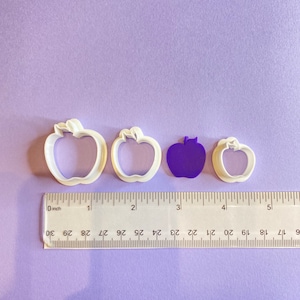 May include: Four white plastic apple-shaped cookie cutters of different sizes, with a purple apple-shaped cookie cutter in the center. The cookie cutters are arranged on a purple surface with a ruler underneath.
