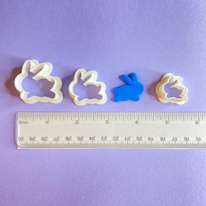 May include: Three white plastic bunny cookie cutters of different sizes, with a blue plastic bunny cookie cutter in the middle. The cookie cutters are arranged in a row on a purple surface with a ruler underneath.