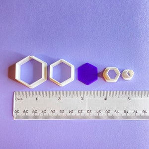 May include: A set of five white plastic cookie cutters in the shape of hexagons, with a purple hexagon in the center. The cutters are arranged in a row from largest to smallest, with a ruler below them for scale.