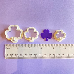 Könnte beinhalten: Drei weiße Plastik-Ausstecher in Form von Kleeblättern, mit einem violetten Kleeblatt-Ausstecher in der Mitte. Die Ausstecher sind in einer Reihe auf einer violetten Oberfläche angeordnet, mit einem Lineal, das 12,7 cm lang ist.
