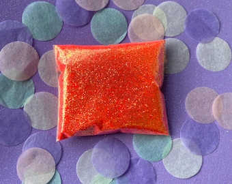 Peut inclure: Un petit sac de paillettes orange entouré de cercles de papier de couleur pastel sur un fond violet.