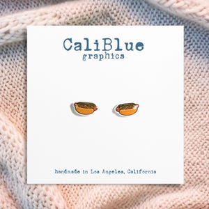May include: A white card with the text "CaliBlue graphics" and two hot dog earrings. The earrings are orange, green, and red. The card also says "handmade in Los Angeles, California."