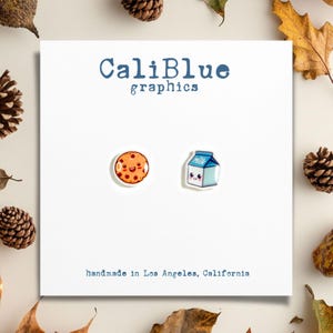 May include: A pair of earrings featuring a cartoon cookie and a cartoon milk carton. The earrings are white with colourful details. The text "CaliBlue graphics" is at the top of the card and the text "handmade in Los Angeles, California" is at the bottom.