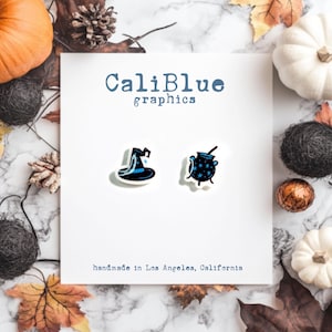 May include: A pair of witch-themed earrings featuring a black witch hat with a blue brim and a cauldron with blue stars. The earrings are on a white card with the text "CaliBlue graphics" and "handmade in Los Angeles, California".