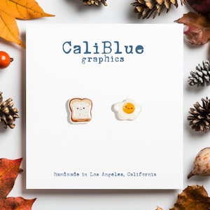 May include: A pair of handmade earrings featuring a smiling fried egg and a slice of toast. The earrings are white with brown and yellow details. The earrings are made in Los Angeles, California. CaliBlue graphics.