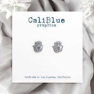 May include: A pair of white hamsa hand earrings with a black evil eye design. The earrings are handmade in Los Angeles, California. CaliBlue graphics.