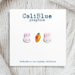 May include: A set of three white bunny and carrot earrings. The earrings are made of polymer clay and are handmade in Los Angeles, California. CaliBlue graphics.