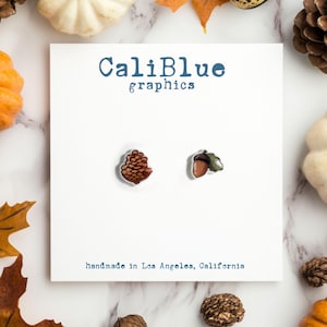 May include: A pair of earrings featuring a brown acorn with a green leaf and a brown pine cone. The earrings are on a white card with the text "CaliBlue graphics" and "handmade in Los Angeles, California".