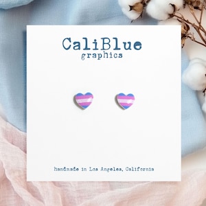 Op de afbeelding: Een paar hartvormige oorbellen met het ontwerp van de transgender pride vlag. De oorbellen zijn roze, blauw en wit. De kaart leest "CaliBlue graphics" en "handmade in Los Angeles, California."