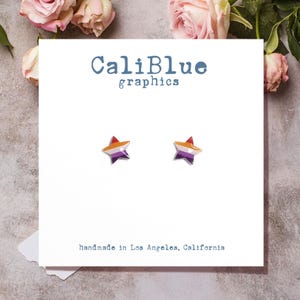 May include: A pair of star-shaped earrings with orange, purple, and silver accents. The earrings are displayed on a white card with the text "CaliBlue graphics" and "handmade in Los Angeles, California".