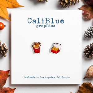 May include: A white card with the text "CaliBlue graphics" and "handmade in Los Angeles, California".  Two pairs of earrings are shown on the card. One pair is a red and white box with yellow french fries. The other pair is a red and white box with yellow chicken nuggets.