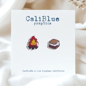 Könnte beinhalten: Eine weiße Karte mit dem Text "CaliBlue graphics" und "handmade in Los Angeles, California". Die Karte zeigt zwei kleine, farbenfrohe Illustrationen: ein Lagerfeuer mit einem lächelnden Gesicht und ein S'more mit einem lächelnden Gesicht.