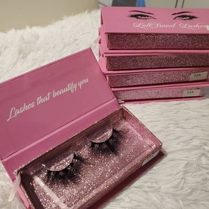 May include: Pink eyelash packaging with glitter and the text "Lash that beautify you" on the lid. The packaging is open to reveal a pair of black false eyelashes.