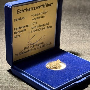 May include: A small, metallic, irregular-shaped meteorite fragment displayed inside a blue hinged box. The interior lid features a certificate of authenticity with the text: Echtheitszertifikat, Fundort: Campo Cielo Argentinien, Entdeckung: 1774, Herkunft: Asteroidengürtel, Alter: 4.500.000.000 Jahre.