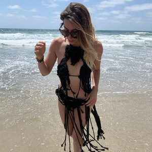 Op de afbeelding: Een vrouw draagt een zwart gehaakt bikini-topje en rok met franjes. Ze staat op een zandstrand met de oceaan op de achtergrond.