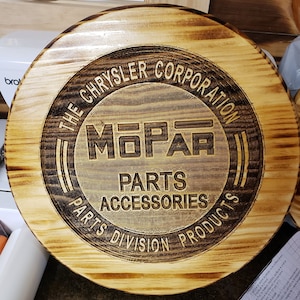 Könnte beinhalten: Runder Holzschild mit dem Mopar-Logo der Chrysler Corporation, mit dem Text "PARTS ACCESSORIES PRODUCTS". Das Schild hat eine hellbraune Oberfläche mit dunkleren, verbrannten Kanten und Schriftzügen.
